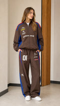 Front full view of woman wearing brown retro racing graphic two-piece tracksuit set with blue and red accents